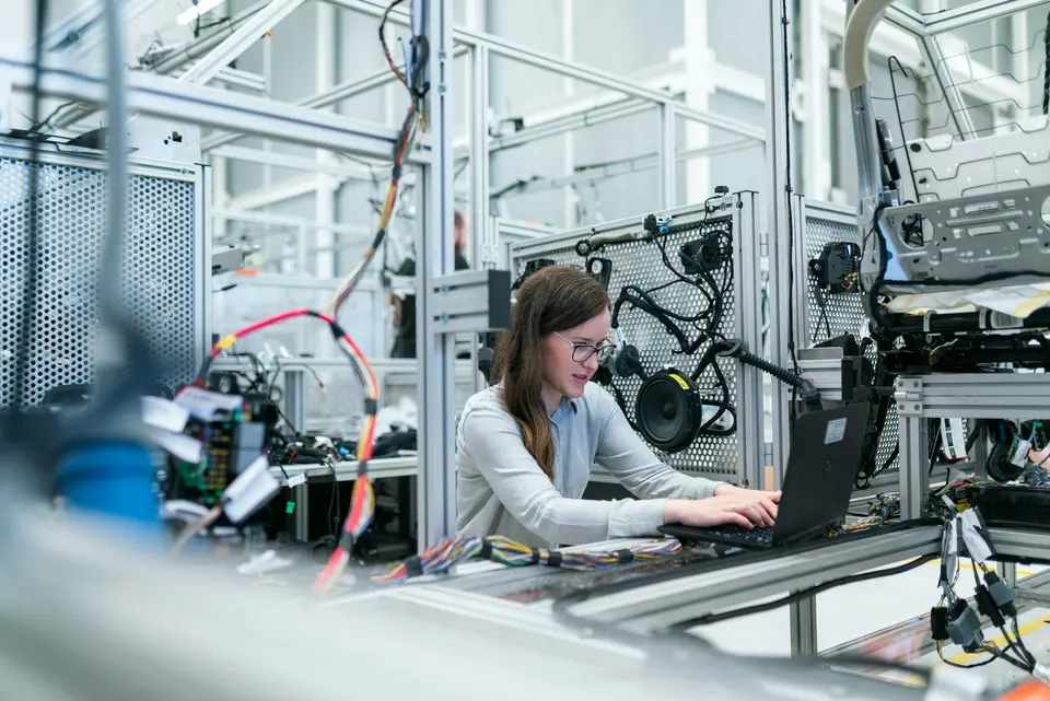 A worker in safety glasses using a laptop beside an automated assembly line.