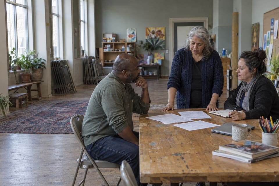 Three adults reviewing papers around a worn wooden table in a community arts room.